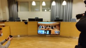 Medierna följer med rättegången via videolänk. Rättegångssal på videoskärm.