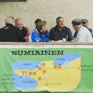 Ryhmä miehiä juo kahvia ja juttelee ulkoterassilla. Terassin kaiteeseen kiinnitetty kartta Sumiaisten kylästä.