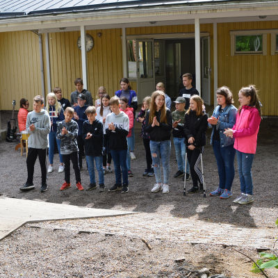 En grupp elever står framför en skolbyggnad. De klappar händer och sjunger.