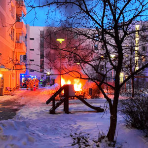 Personal från räddningsverket står i grupp bredvid en lekstuga som brinner mellan en mängd bostadshus.