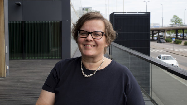 En kvinna i medelåldern med kort hår, glasögon och svart tröja står på en större balkong med trägolv. I bakgrunden syns en trafikerad väg och en bro.