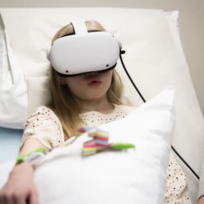Vaaleahiuksinen pikkutyttö pilkullisessa paidassa virtuaalilasit päässä pää valkoisella sairaalatyynyllä.