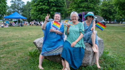 Tre damer sitter på en sten och viftar med små regnbågsflaggor.
