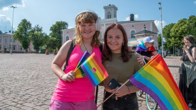 Två unga flickor står på Lovisa torg med regnbågsflaggor. De tittar in i kameran och ler.