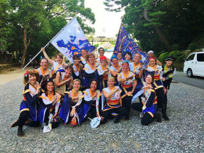 Ett yosakoi danslag i Japan. Alla är kvinnor och de bär vita och blåa dräkter. Två av dem håller i stora flaggor som också går i vitt och blått. 