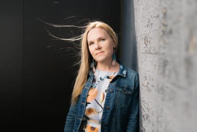 Barnombudsman Elina Pekkarinen står lutad mot en fasad. Hon är klädd i blommig skjorta och jeansjacka. 