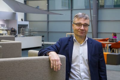 Matti Merisalo i blå kavaj står med armbågen stödd mot en soffgrupp.