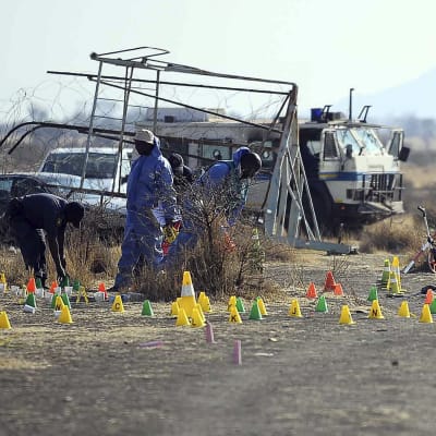 Några personer i blå overaller utför undersökningar efter massakern i Marikana. Gula, gröna och orange markeringar på marken. Några fordon i bakgrunden. 