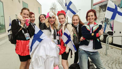 Krista iklädd brudklänning tillsammans med sina dansare på väg mot finalen i Eurovisionen. De poserar med Finlands-flaggor och ler rakt in i kameran.