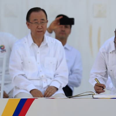Ban Ki-moon istuu vieressä, kun Rodrigo Londono Echeverri allekirjoittaa sopimusta. Taustalla näkyy muita ihmisiä. Kaikki ovat pukeutuneet valkoiseen.
