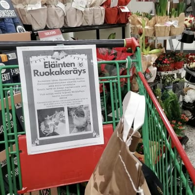 Ostoskärryyn kiinnitetty keräyslappu kaupassa. Lapussa kerrotaan eläinten ruokakeräyksestä ja siinä on kuvia kissoista.