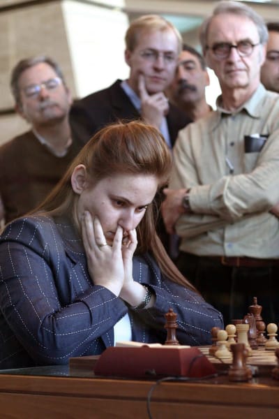 En kvinna spelar schack. Flera män står runt omkring henne och tittar på.