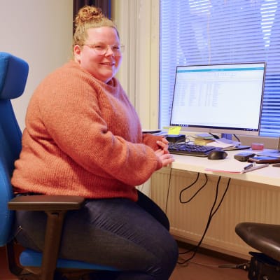 En kvinna i orange tröja sitter framför ett skrivbord med två datorskärmar. Hon ler och tittar in i kameran. Bakom henne syns en bäddad säng.