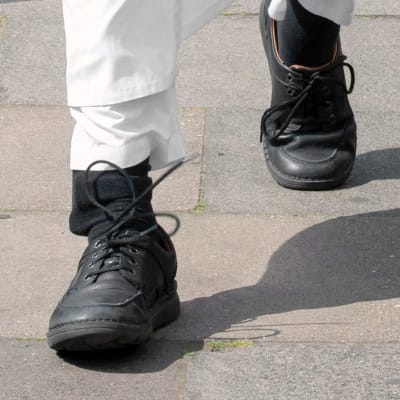 Fötter med svarta skor och svarta strumpor. Under syns en fotboja.
