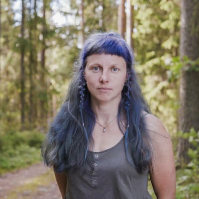 Hantverkskonstnären Elena Bondar i närbild. I bakgrunden en tät granskog. Elena Bondar har långt, blåfärgat hår med pannlugg.