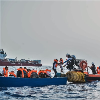 Siirtolaisia autetaan sinisestä veneestä oranssireunaiseen kumiveneeseen. Sukelluspukuinen mies ottaa juuri vastaan lasta. Taustalla näkyy punavalkoinen Ocean Viking -alus.