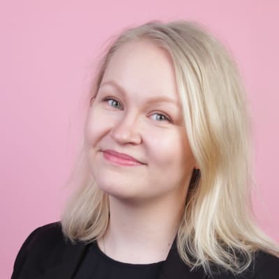 En kvinna med ljust axellångt hår, svart skjorta och svart kavaj tittar in i kameran. Bakgrunden är rosa.