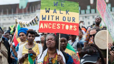 En kvinna går längst fram i en marsch med en skylt där det på engelska står "We walk with our ancestors"