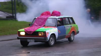 Lada saapuu savuverhon takaa. Siinä on suuri vaaleanpunainen rusetti katolla.