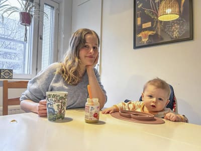 En kvinna och ett spädbarn sitter vid ett bord. De ser båda in i kameran med allvarlig min. På bordet finns mat.
