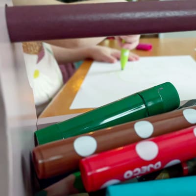 Lapsen värikyniä työkalupakissa. Taustalla lapsen käsi piirtää tussilla paperiin.
