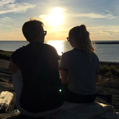 En mamma och en tonårsdotter sitter på klippa i solnedgången och pratar med varandra.