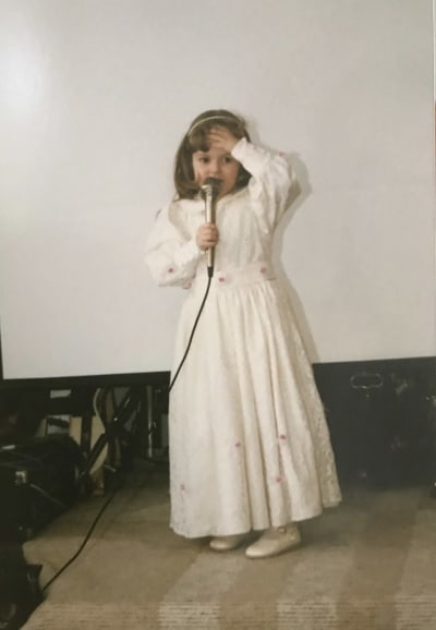 Muusikko Ellinoora pikkutyttönä pitkässä juhlamekossa laulamassa mikrofoni oikeassa kädessä, vasen käsi otsalla.