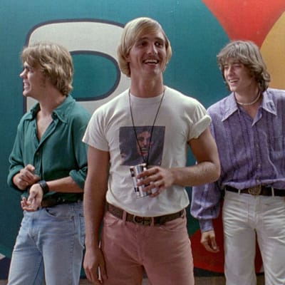Neljä nuorta poikaa tai miestä 1970-luvun tyylisissä vaatteissa seisoo rennosti hymyillen, etualan tyypillä kädessään oluttölkki, taustalla kirjava seinä, kuva elokuvasta Surutta? Sekaisin.