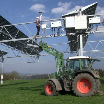 En traktor på en tysk åker med solpaneler högt ovanför marken. En man håller på med underhållsarbete. 