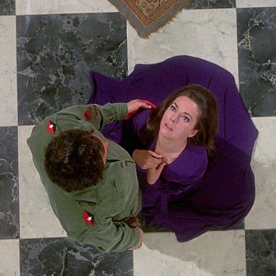 Suoraan ylhäältä otetussa kuvassa kaunis tummahiuksinen nainen violetissa mekossa on kaatumassa shakkiruutukuvioiselle lattialle, vihreässä kenttäunivormussa oleva mies pitelee veristä kättään naisen olkapäällä. Kuva Hitchcockin elokuvasta Topaz.