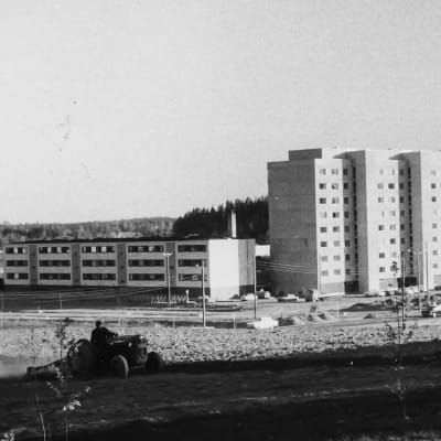 Traktor på åker framför förortshöghus.