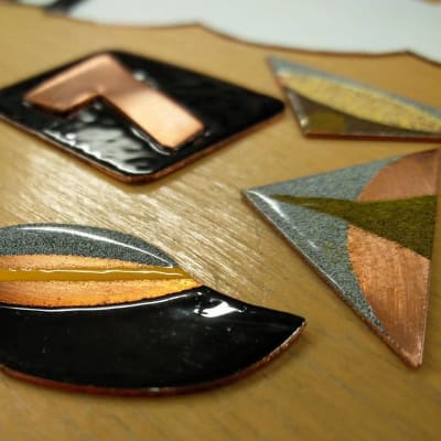 Små kopparbitar i olika former som emaljerats med olika färger på en brun bordsskiva.