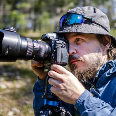 Luontokuvaaja Antti Haataja metsässä järjestelmäkameran kanssa. Kuvan kulmassa KC-logo.