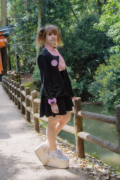 En kvinna klädd i kavaj, skjorta, kort kjol och skor med hög platåsula. Hon står vid en bro och på höger sida syns lite vatten.
