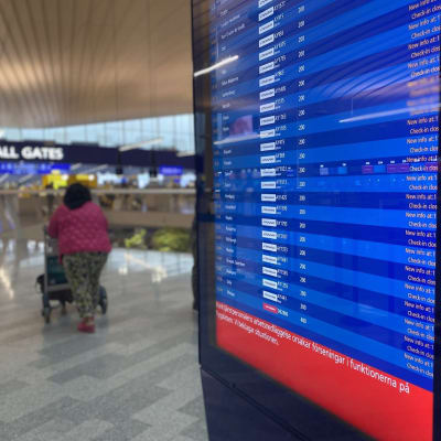 Helsinki-Vantaan lentoaseman infotaulu lähtevistä ja saapuvista lennoista, taustalla nainen työntää matkalaukkukärryä.