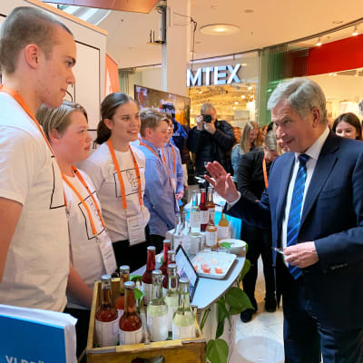 Till vänster synns tre umgdomar som står bakom ett bord med läskedrycker på. Till höger synns president Sauli Niinistö som pratar med dem.