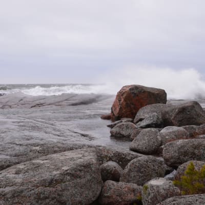 En strand med stora släta stenar. Stora vågor slår mot stenarna och himlen är grå.