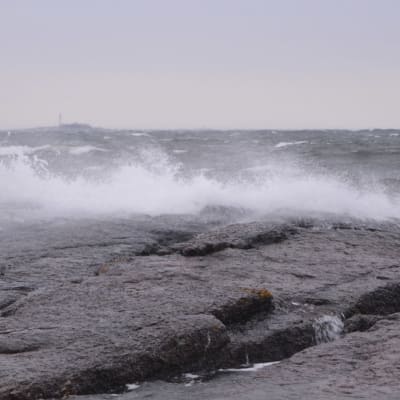 Meren aallot lyövät vaahdoten rantakallioon. Taivas on harmaa ja keli myrskyinen.