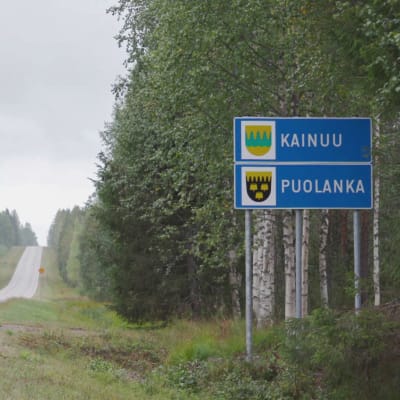 Tyhjä maantie kesällä, tien vieressä kyltti, jossa lukee 'Kainuu' ja 'Puolanka'.