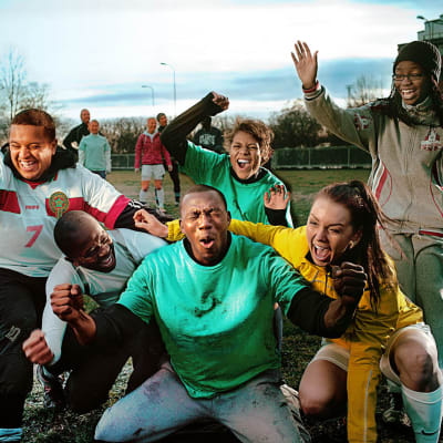 kuusihenkinen ryhmä jalkapallon harrastelijapelaajaa iloitsee etualalla, takana seisoo toinen joukkue paikoillaan