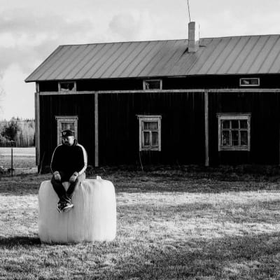 Joel sitter på en höbale framför ett torp. Bilden är svartvit.