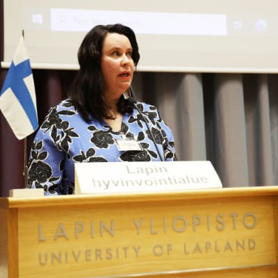 Johanna Ojala-Niemelä i blåblommig klänning håller tal i en talarstol.