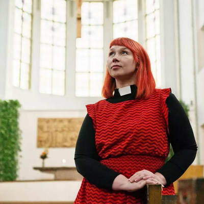 Pastor Marjaana Toiviainen i röd klänning. Bilden är tagen lätt underifrån och Marjaana har blicken vänd uppåt.