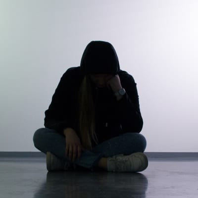 Mustaan huppariin pukeutunut nuori istuu pää painuneena jalat ristissä.