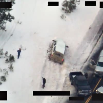 Kuva on otettu ylhäältä. Lumisessa maisemassa kaksi poliisia lähestyy molemmilta suunnilta hattupäistä miestä. Yksi auto on ajanut lumeen tieltä, kolme autoa on tiellä kolmion muotoisessa asetelmassa.