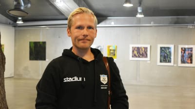 En ung man i svart luvtröja står inne i Konstfabriken i Borgå. Bakom honom syns grå väggar och tavlor.