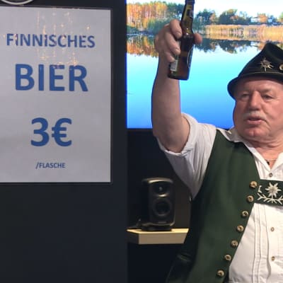En man klädd i tyska kläder skålar framför en skylt med texten Finnishes bier.