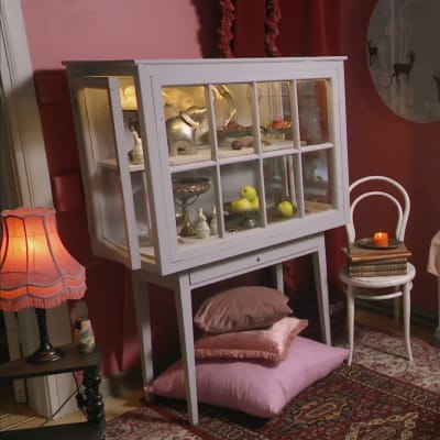 Ett gråmålat vitrinskåp som är gjort av gamla fönster. Skåpet står i ett vardagsrum med rosa väggar.
