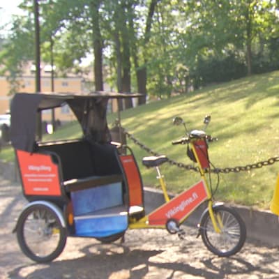 En man med gul skjorta och keps står vid sin cykeltaxi, riksha, vid en park en varm sommardag.