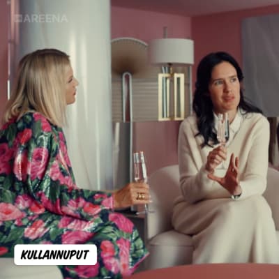 Kaksi naista istuu keskustelemassa vaaleanpunaisella sisustetussa huoneessa shampanjalasit kädessään.